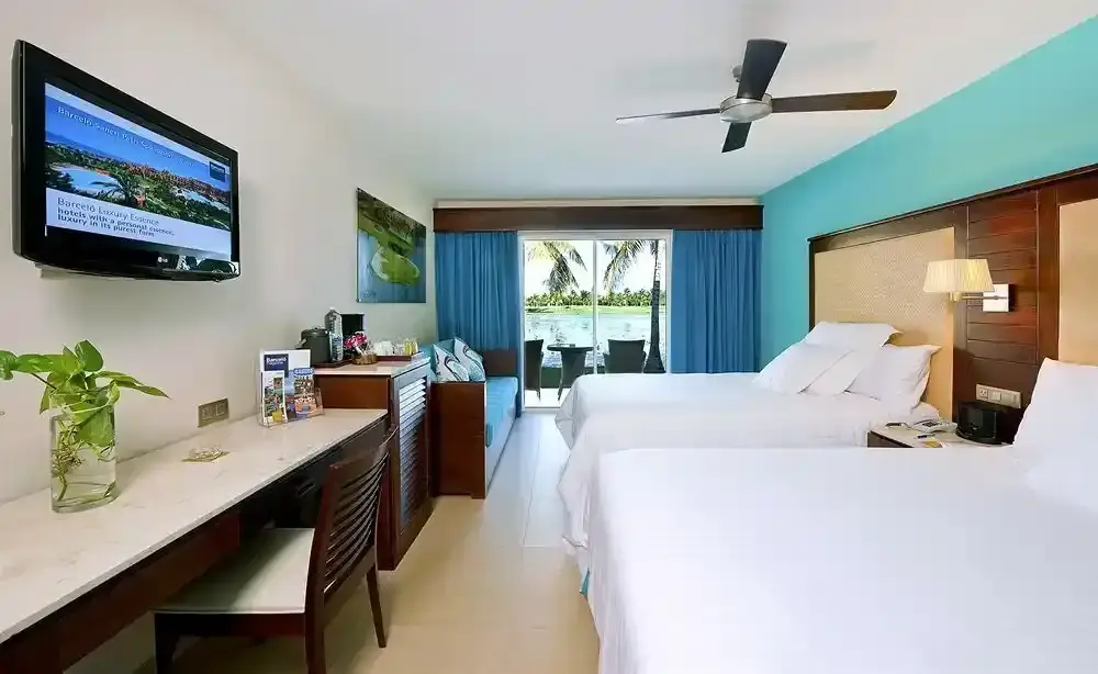 Hotel Room at Barceló Bávaro Palace Punta Cana Modern hotel room at Barceló Bávaro Palace with two double beds, desk, flat-screen TV, and balcony overlooking tropical palm trees and the Caribbean Sea.